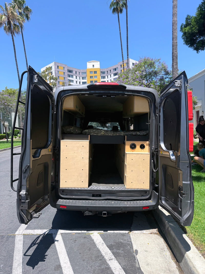 Custom RV rental campervan conversion with open rear doors revealing a bed, wood storage, and speakers. Parked outdoors on a sunny day. — photo 4