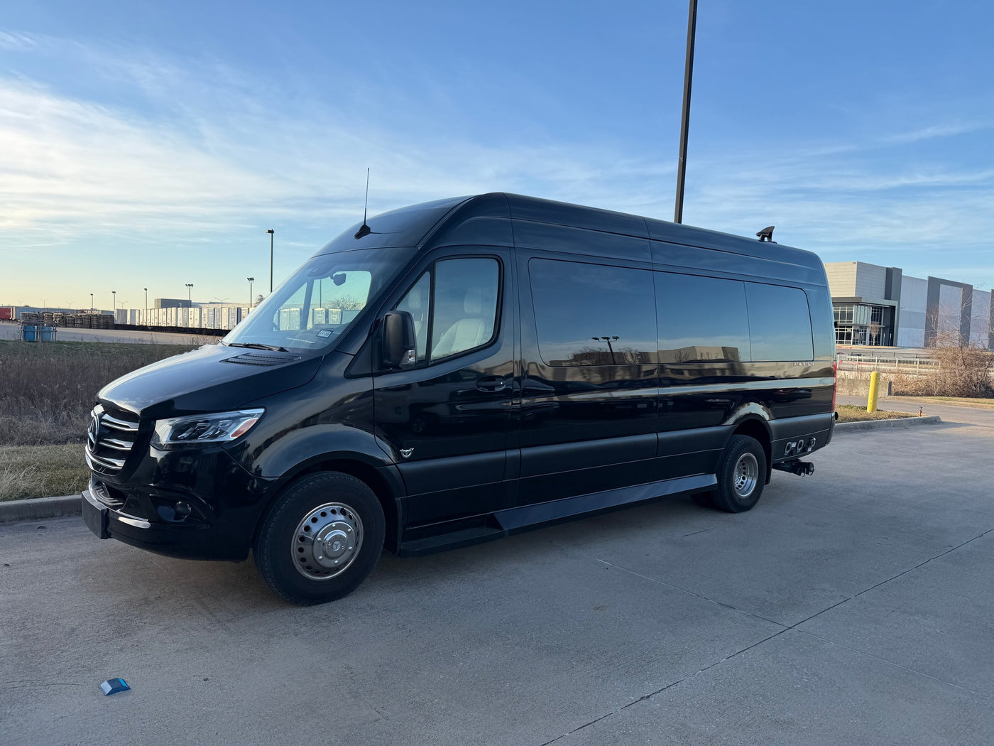 A black Mercedes-Benz Sprinter luxury van, ideal for rv rental, parked in a lot under a bright blue sky with industrial buildings behind it. — photo 6