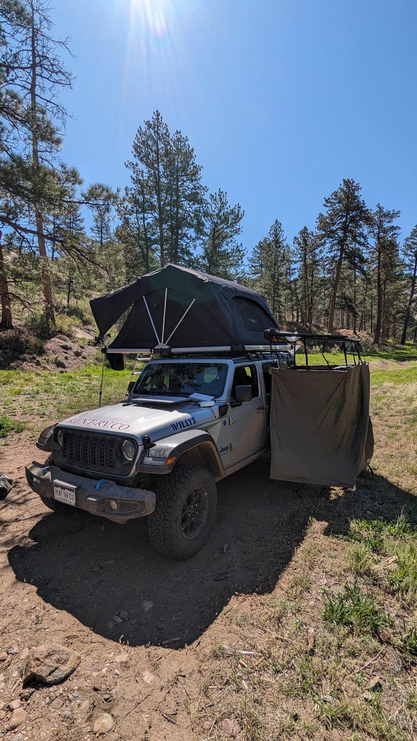 Jeep RV rental with rooftop tent & privacy screen set up for overlanding and camping in a sunny forest.