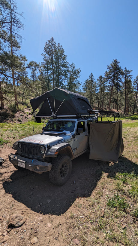Jeep RV rental with rooftop tent & privacy screen set up for overlanding and camping in a sunny forest.
