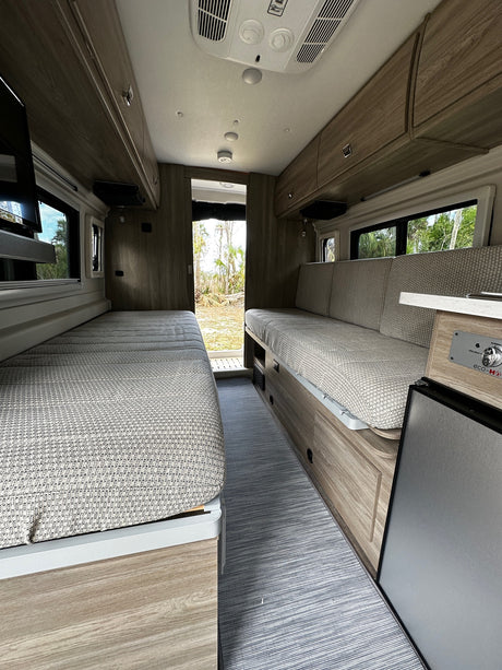 Inside an RV rental: a compact, modern interior with two textured benches convertible to beds, ample storage, and small fridge.
