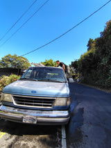 Man washing blue Ford van, prepped as an rv rental, with a ladder on roof under clear skies.