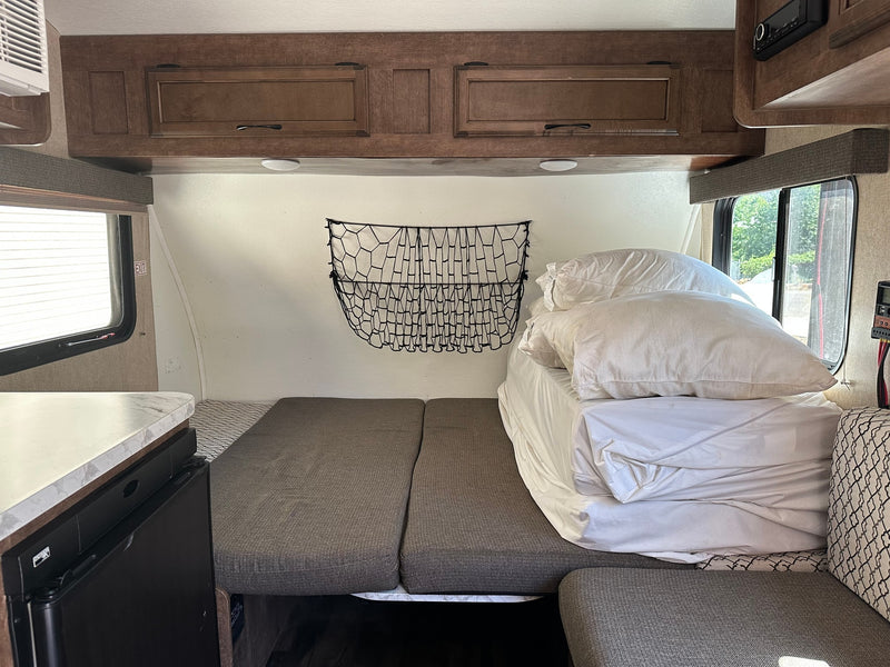 RV rental interior features a neatly made bed with white linens, wooden overhead cabinets, and a side window, ready for travel. — photo 8