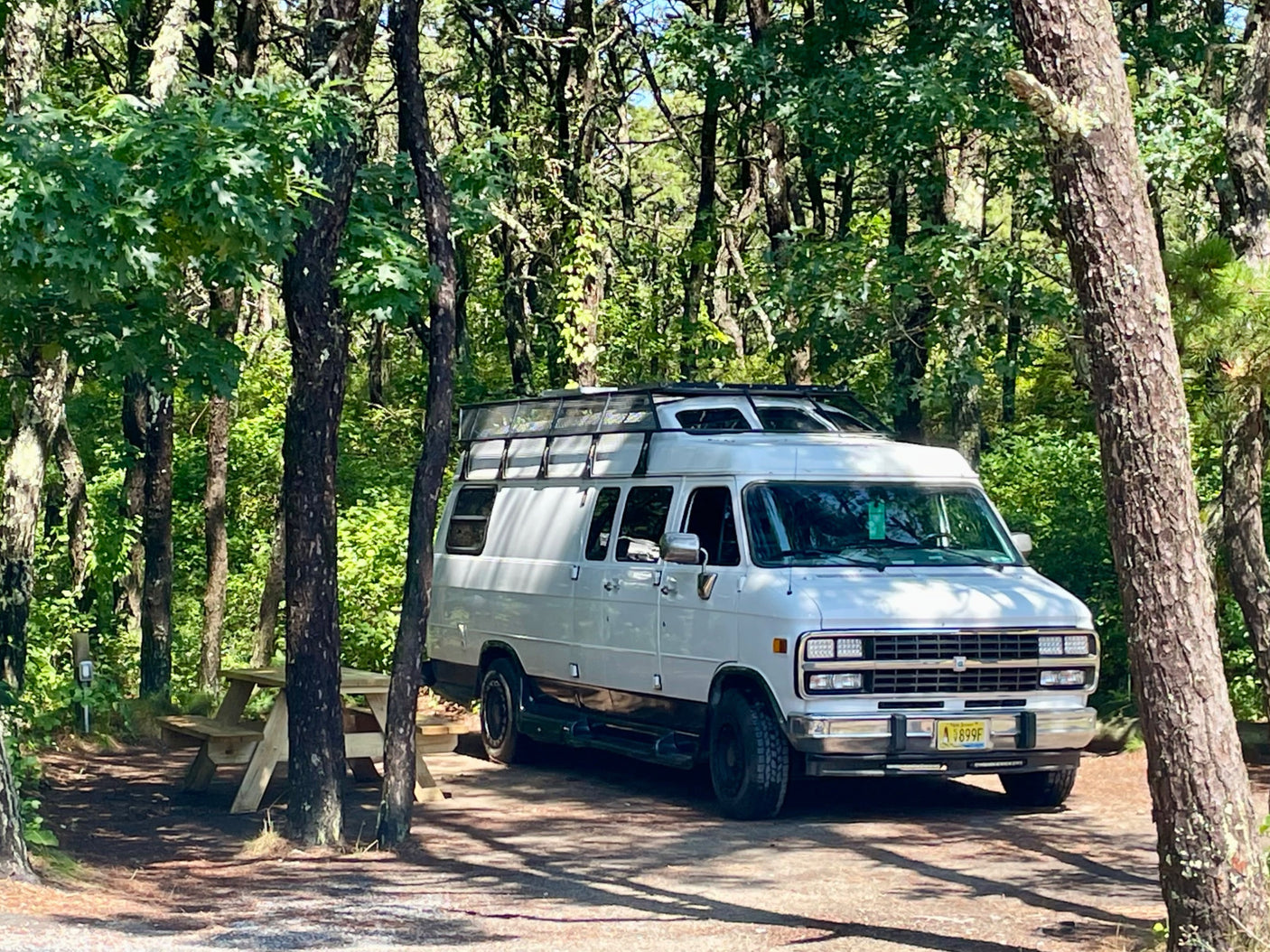 White conversion RV rental parked at a peaceful wooded campsite with a picnic table. Experience nature's serenity.