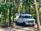 White conversion RV rental parked at a peaceful wooded campsite with a picnic table. Experience nature's serenity.