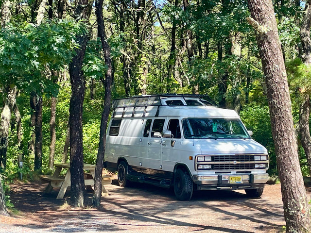 White conversion RV rental parked at a peaceful wooded campsite with a picnic table. Experience nature's serenity.