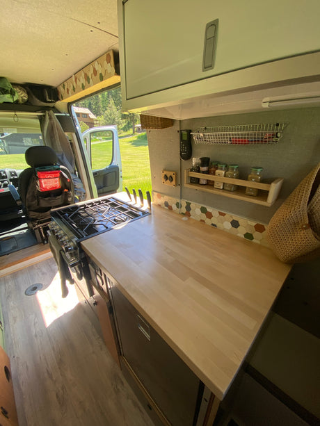 RV rental kitchen: stove, light wood counter, spice rack, & hexagonal tile backsplash. Open door shows a grassy view.