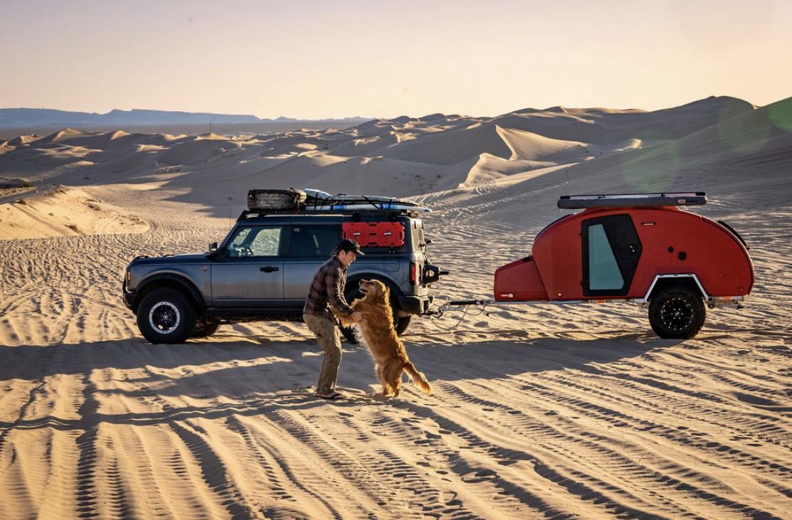 Off-roading in style! A man plays with his dog by a grey Bronco SUV and red teardrop camper in sand dunes. Explore with an RV rental.|Red escapod teardrop RV rental with a mountain bike mounted on top