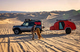 Off-roading in style! A man plays with his dog by a grey Bronco SUV and red teardrop camper in sand dunes. Explore with an RV rental.