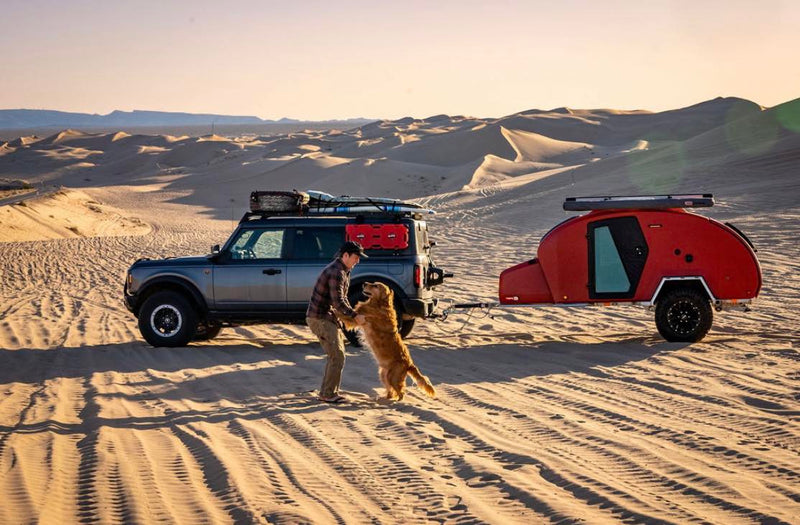 Off-roading in style! A man plays with his dog by a grey Bronco SUV and red teardrop camper in sand dunes. Explore with an RV rental.|Red escapod teardrop RV rental with a mountain bike mounted on top — photo 1