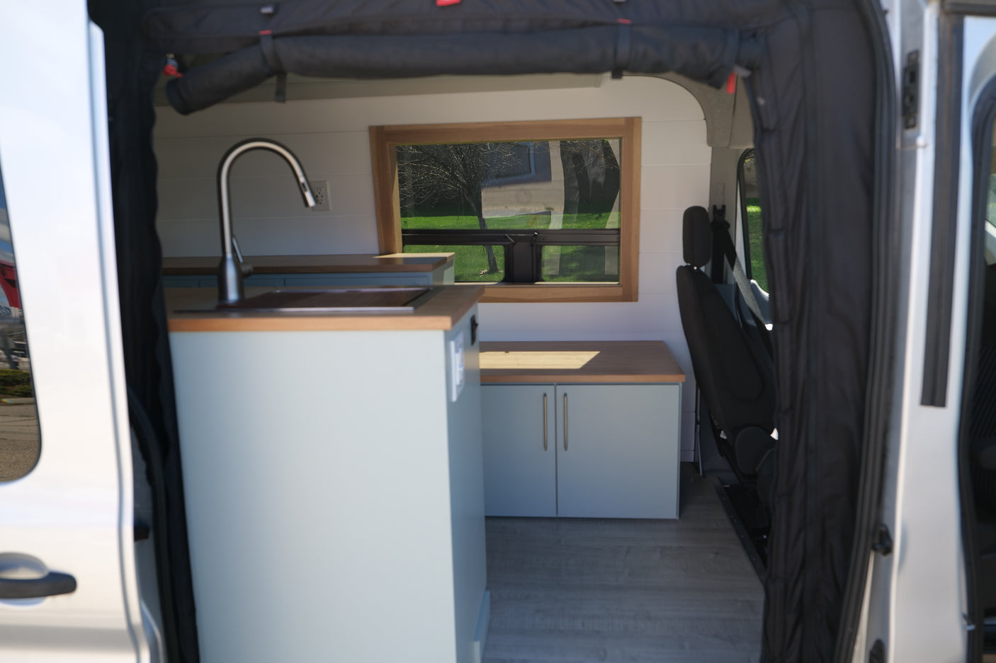 Inside a spacious RV rental camper van: light blue kitchenette with sink, wood countertops, and a window overlooking nature.