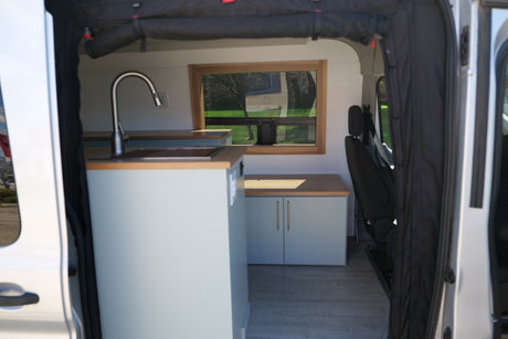 Inside a spacious RV rental camper van: light blue kitchenette with sink, wood countertops, and a window overlooking nature.