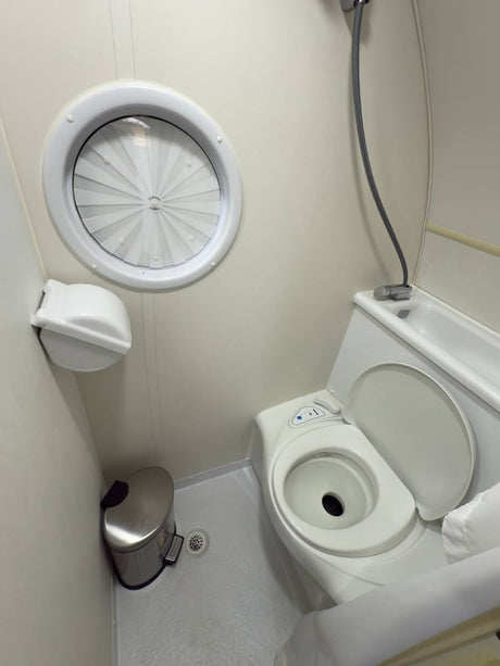 Inside an RV rental bathroom, featuring a compact toilet, shower head, vent, and floor drain. Essential facilities for travel.