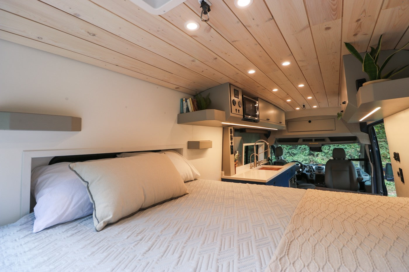 Inside a spacious RV rental: a bright interior with a comfy bed, functional kitchen, and driver's cabin visible through to nature.