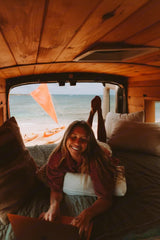 Remote work with an RV rental! Happy woman on laptop inside a wood-paneled van, overlooking a sunny beach & sailboat.