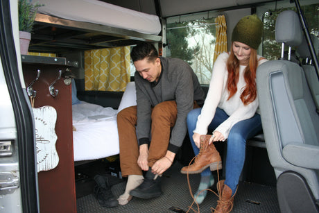 A couple inside an RV rental, putting on shoes. The cozy interior features a bed, storage, and patterned curtains.