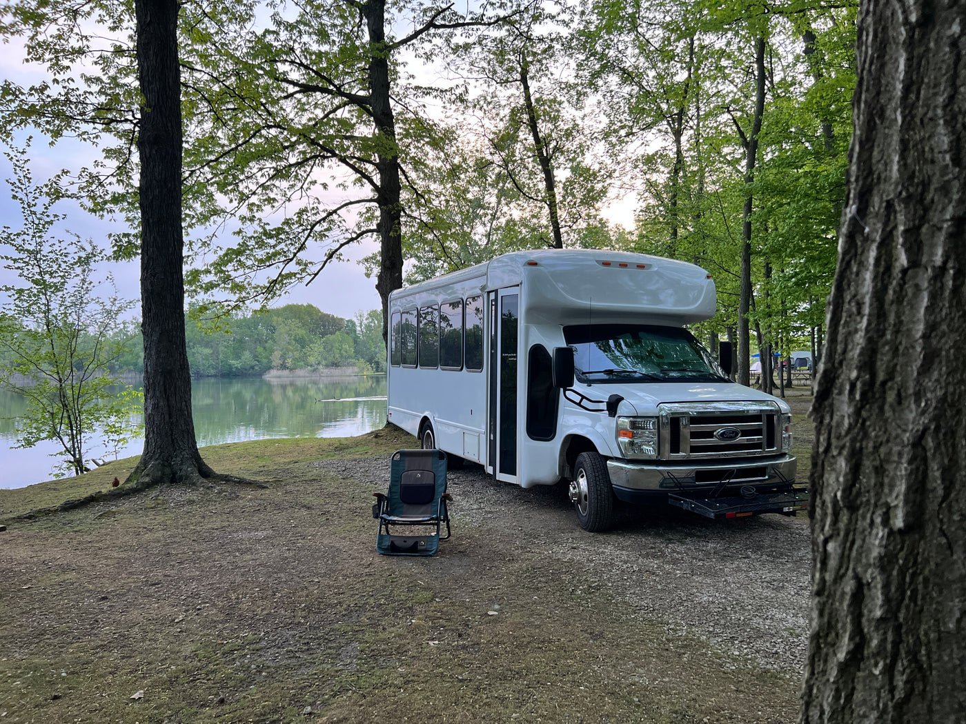Serene lakeside RV rental: a white converted bus is parked under trees, with a camping chair by the calm water. — photo 5