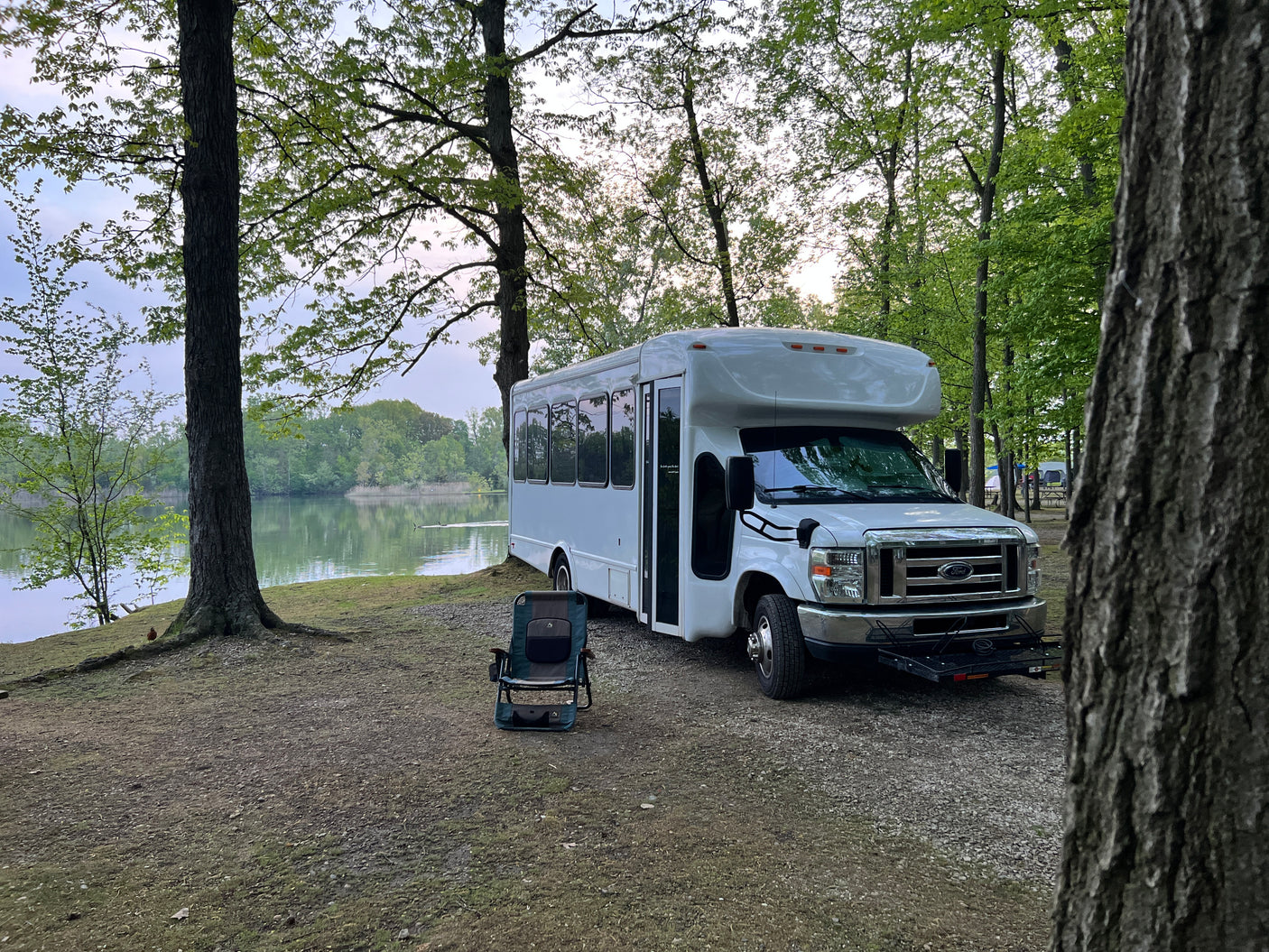 Serene lakeside RV rental: a white converted bus is parked under trees, with a camping chair by the calm water.