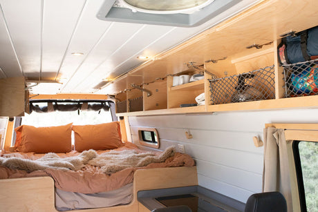 This RV rental's cozy interior features a comfy bed with orange pillows and smart overhead storage for efficient van living.