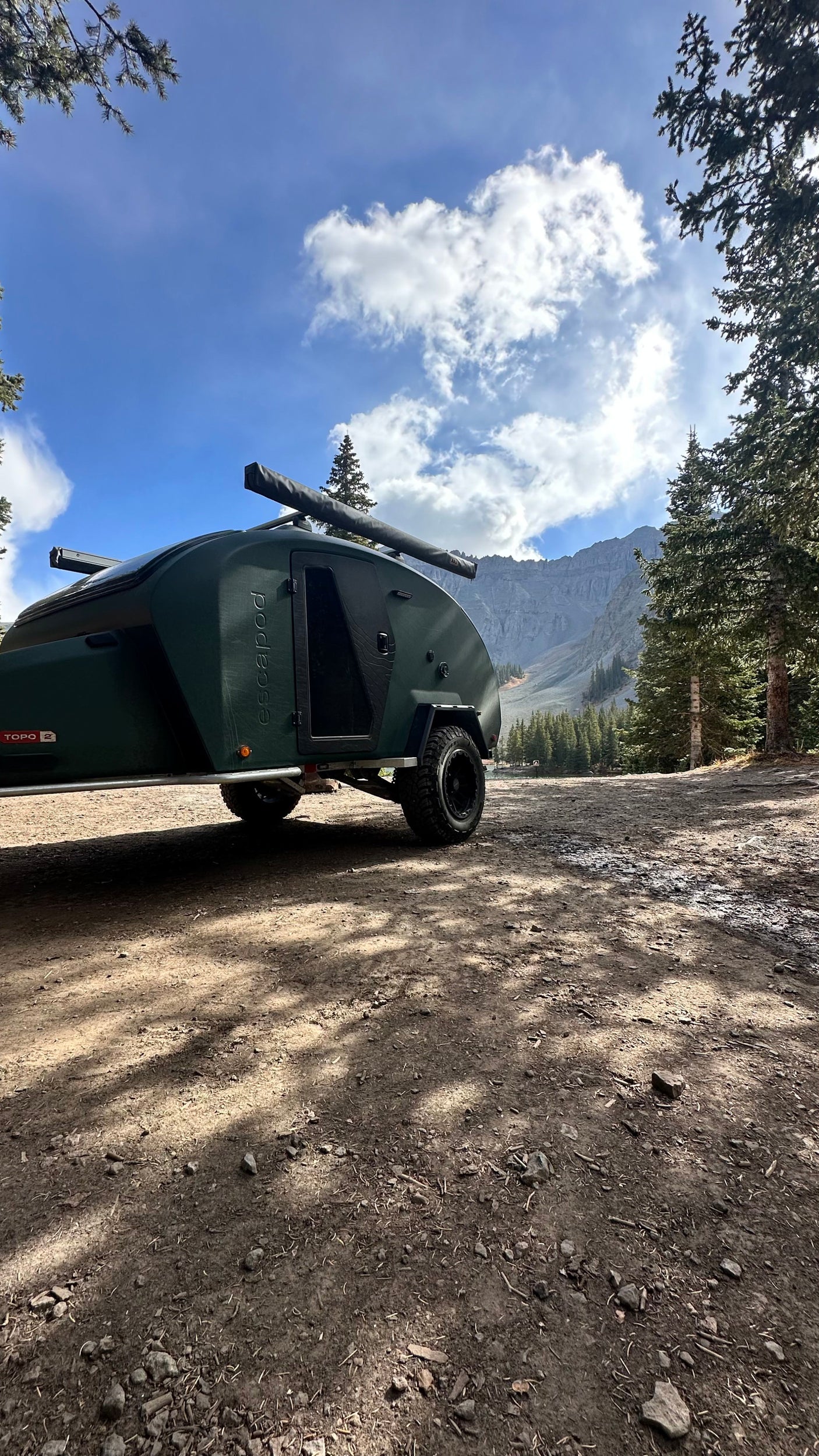 An Escapapod off-road trailer sits on a dirt road near tall pines with mountains behind. Consider this for your next rv rental.