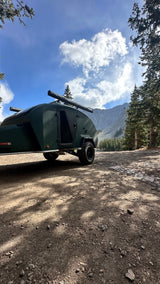 An Escapapod off-road trailer sits on a dirt road near tall pines with mountains behind. Consider this for your next rv rental.