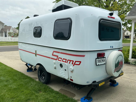 White Scamp compact travel trailer, an ideal RV rental, parked on a driveway with leveling jacks, AC, and rear camera.