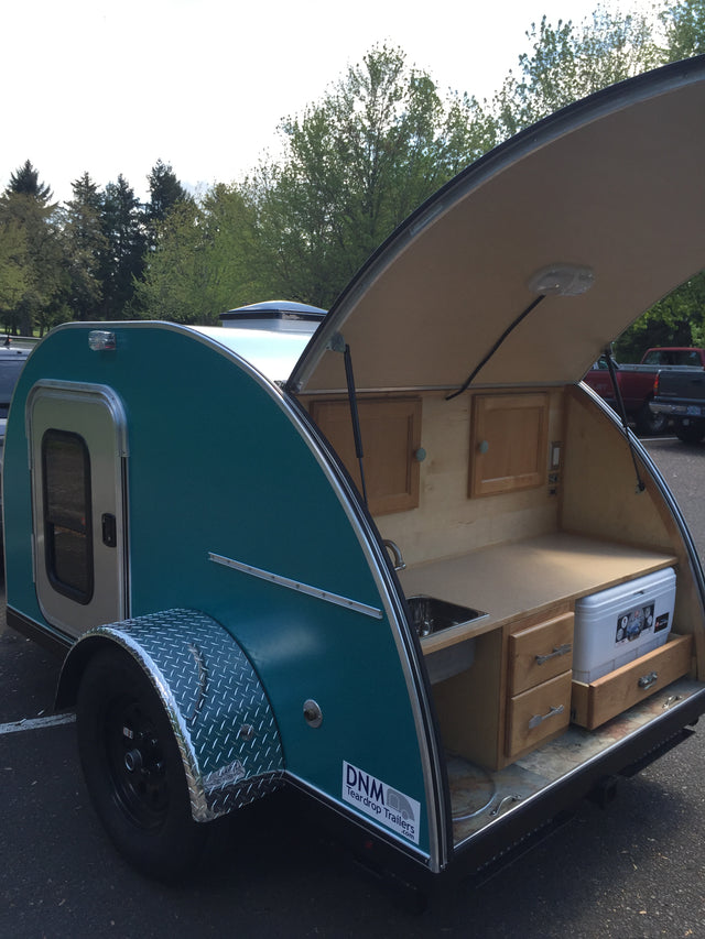 A teal DNM Teardrop Trailer with its rear kitchen open, featuring a cooler and wooden cabinets. Perfect for an rv rental adventure.