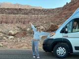 Woman with an RV rental enjoying scenic desert travel. She stands by a silver camper van with rocky cliffs behind.