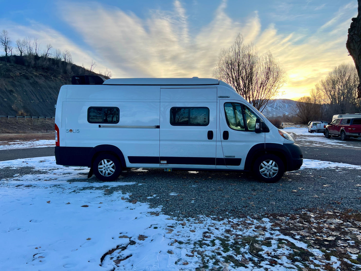White Winnebago Solis camper van for RV rental, parked in a snowy setting with a sunset over mountains.