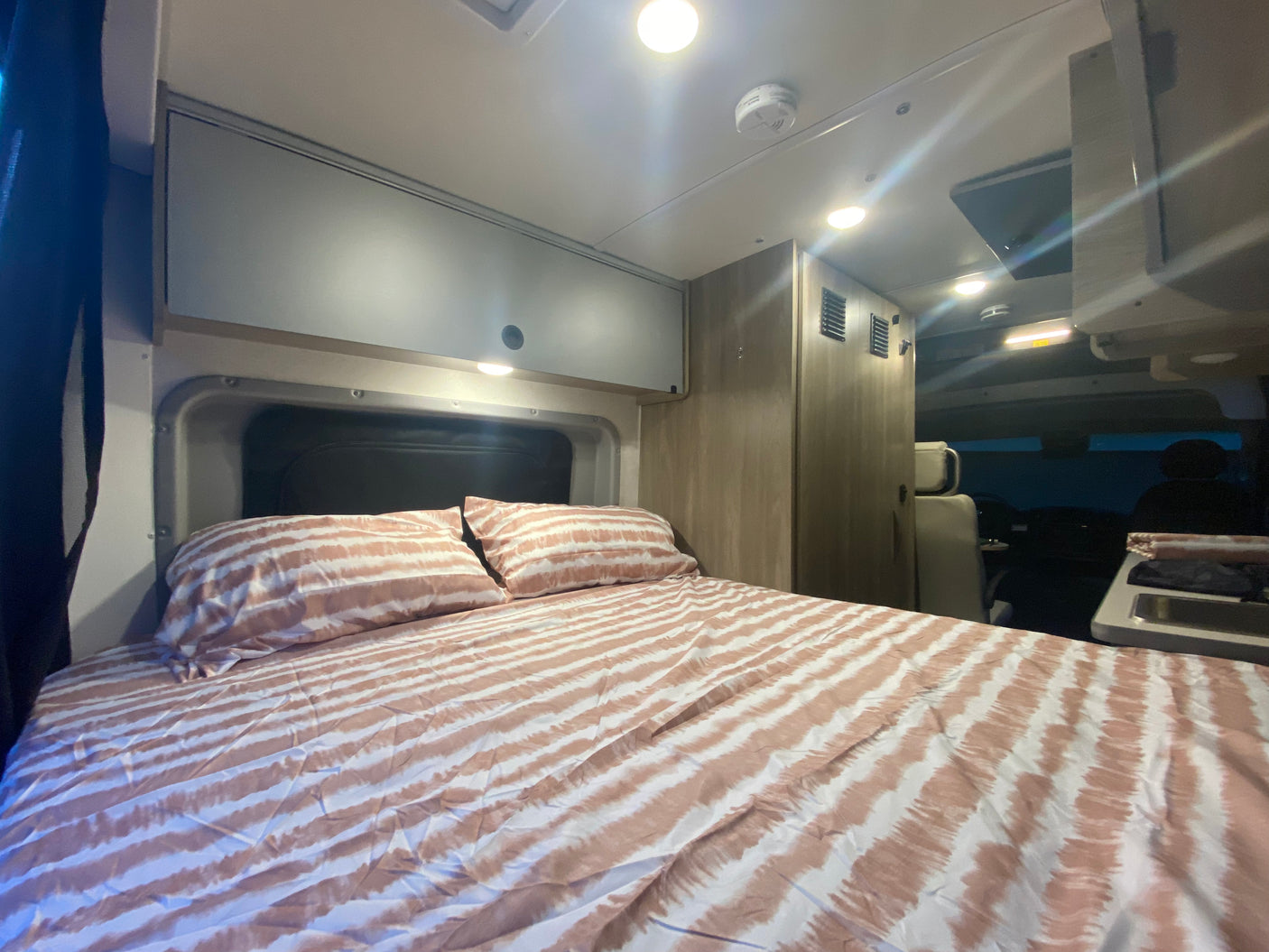 This RV rental bedroom features a bed with striped bedding, overhead cabinets, and a compact kitchenette.