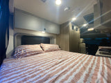This RV rental bedroom features a bed with striped bedding, overhead cabinets, and a compact kitchenette.