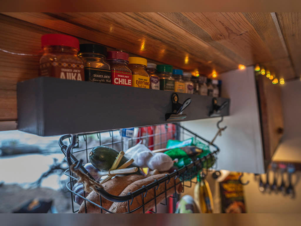 RV rental kitchen: Organized spice shelf with Trader Joe's jars & hanging produce basket. Compact setup.