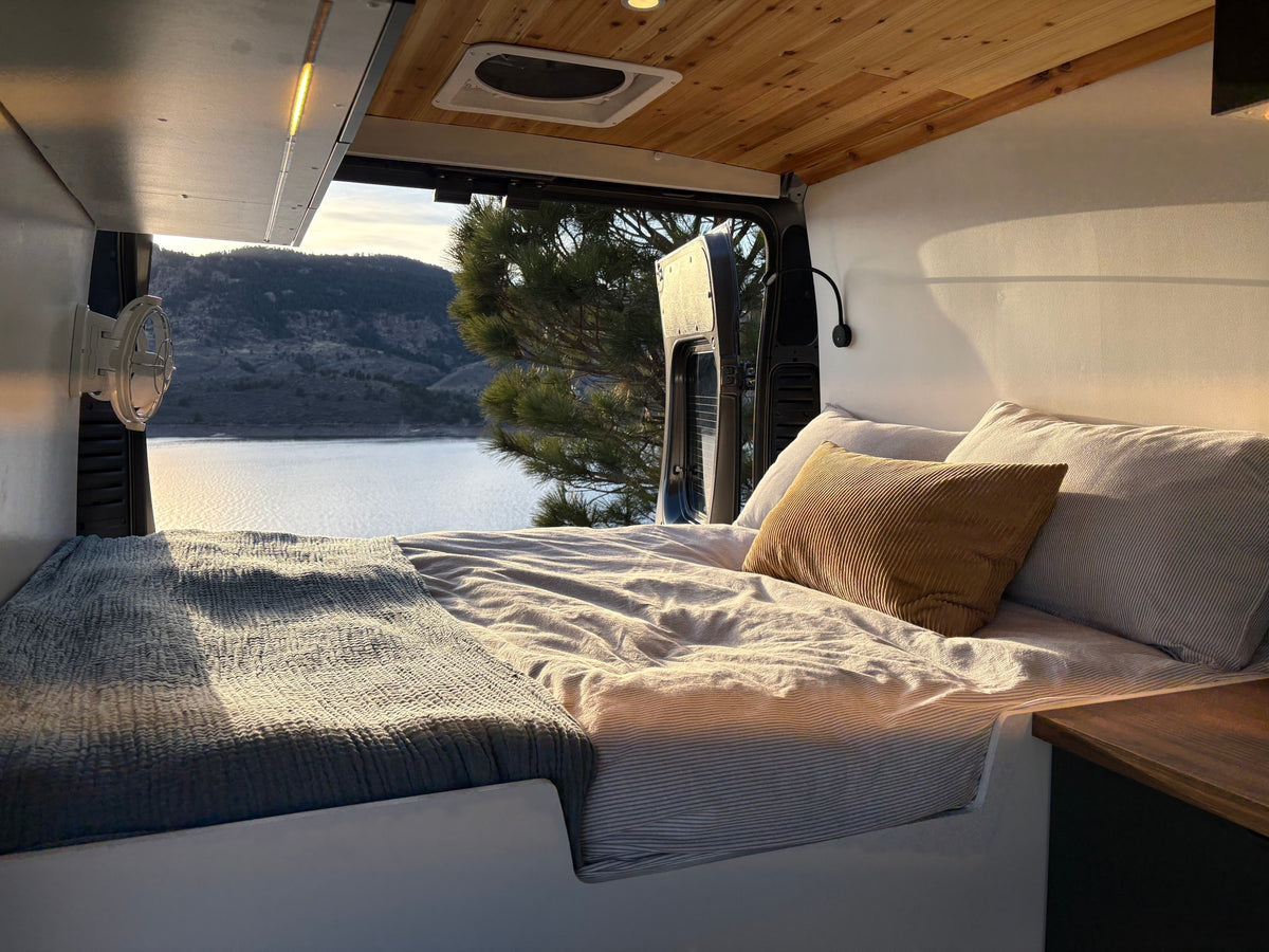 Serene RV rental interior: A comfy bed with a view of a tranquil lake and pine tree through the open van doors. — photo 2