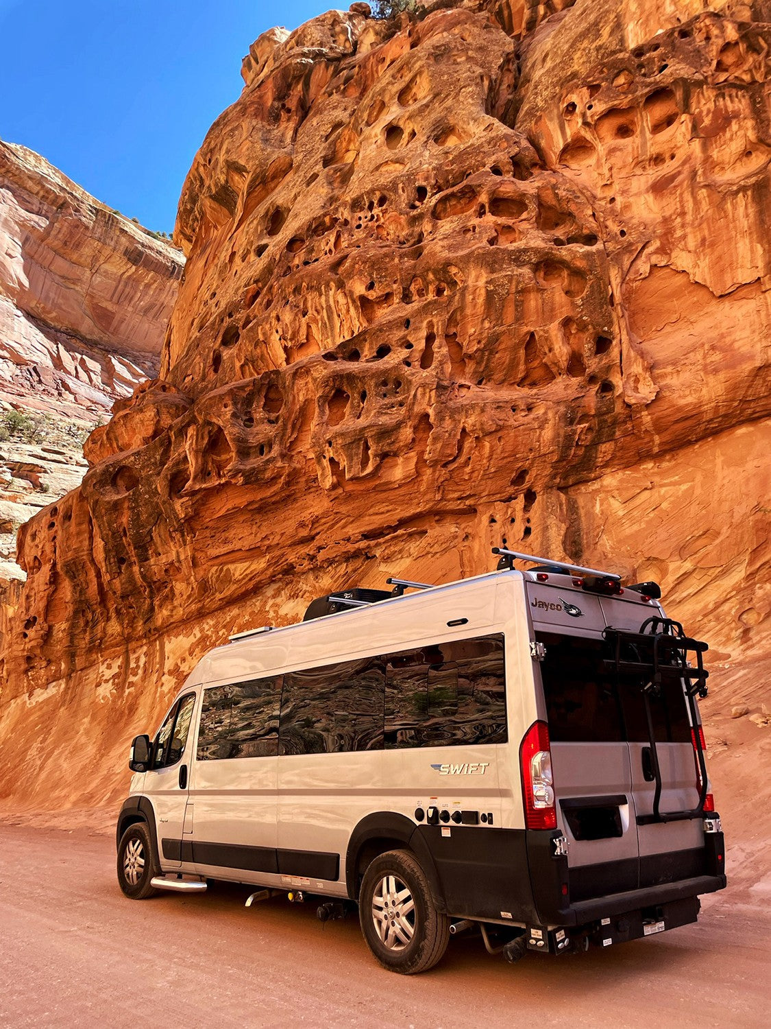 An RV rental, a Jayco Swift campervan, parked alongside towering red rock canyon walls under a clear blue sky.