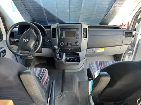 RV rental interior: View of the driver's cockpit, dashboard, and front seats with patterned covers. Sunshade on windshield.
