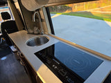RV rental kitchen: modern induction cooktop, sink, and 'Mickey happy place' decor on a light counter. Sunny drive outside.