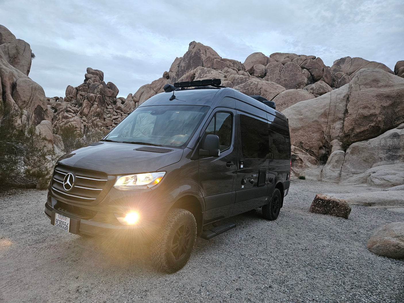 This dark RV rental van, headlights on, is ready for adventure in Joshua Tree's rocky desert.
