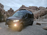 This dark RV rental van, headlights on, is ready for adventure in Joshua Tree's rocky desert.