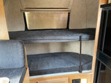RV rental interior featuring stacked bunk beds with grey cushions. A window is above the top bunk, with wooden trim and power outlets below.