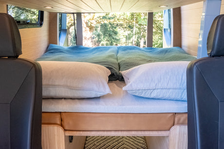 Inside an RV rental: a cozy bed with striped pillows & a blue-green blanket, set in a wood-paneled camper van.