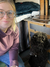 A woman in glasses smiles with her dog inside a cozy RV rental, showing wooden cabinetry and bed area.