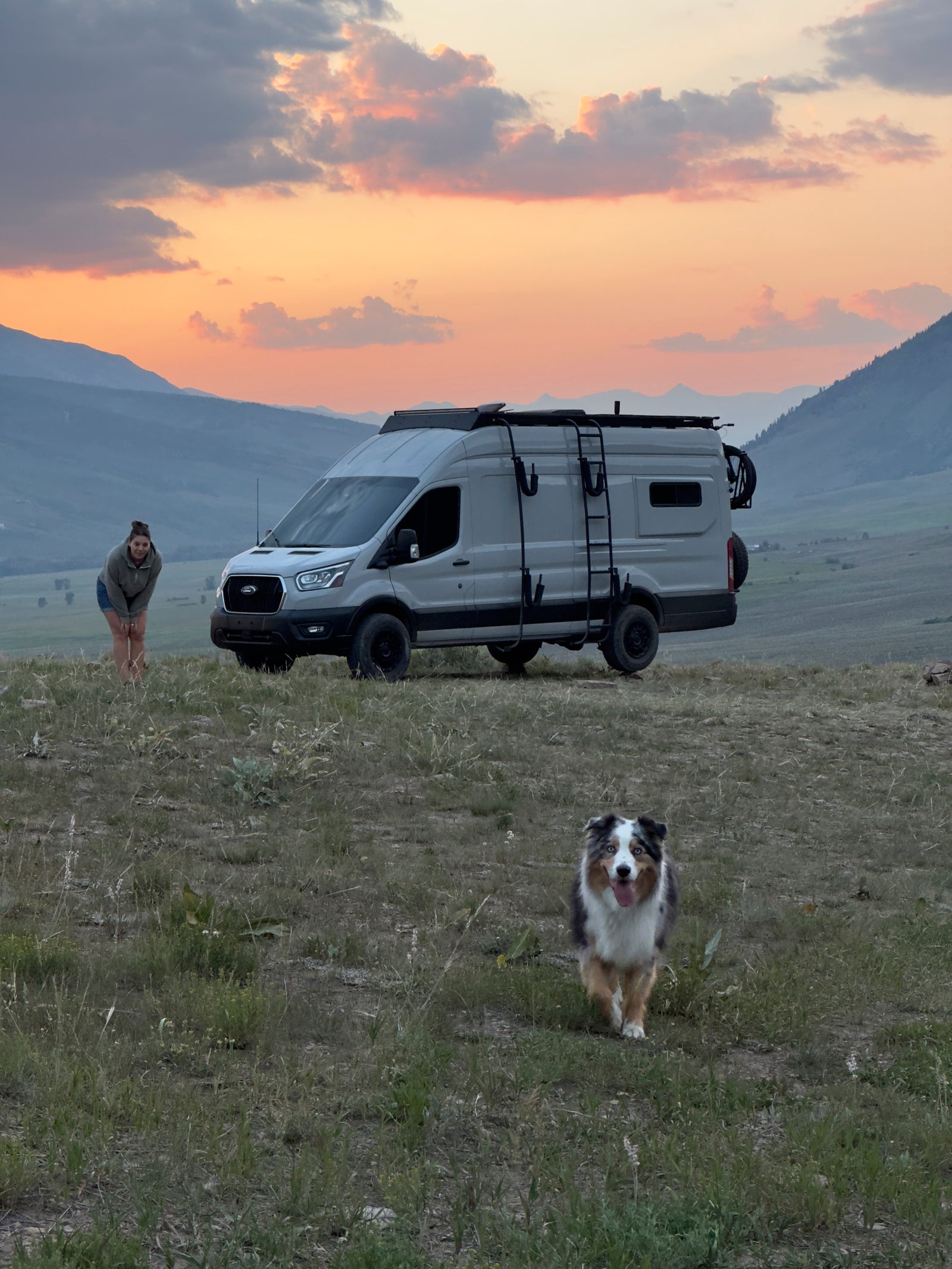 Embrace van life! An RV rental, a woman, and her happy dog at sunset in the mountains. Perfect for outdoor adventures.