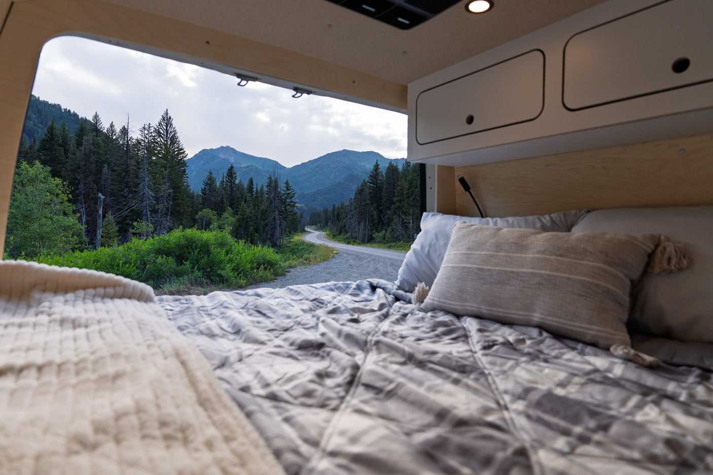 Cozy RV rental bed with an open view of a winding mountain road, lush forest, and peaks.