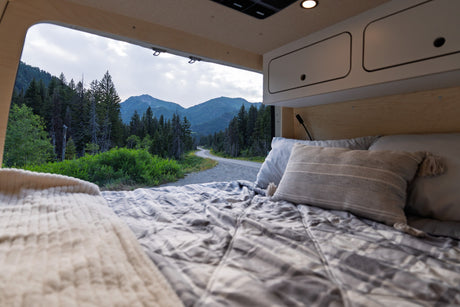 Cozy RV rental bed with an open view of a winding mountain road, lush forest, and peaks.