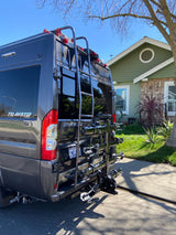 Rear view of a grey Winnebago Travato RV rental, featuring a ladder and multi-bike rack, parked near a house.