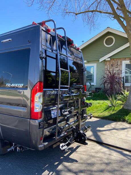 Rear view of a grey Winnebago Travato RV rental, featuring a ladder and multi-bike rack, parked near a house.