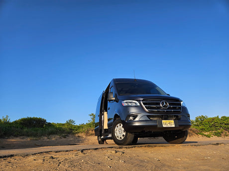 A dark grey Mercedes Sprinter van on a sandy road, driver's door open. Great RV rental for clear sky adventures.