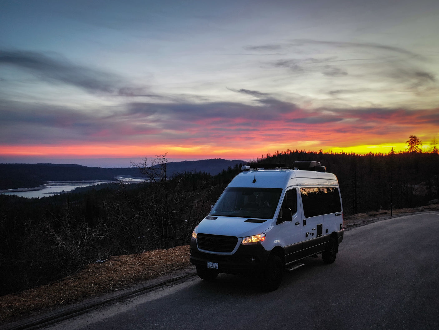 White camper van on a road at sunset, overlooking vibrant pink/orange skies over mountains and a lake – perfect rv rental view!