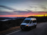 White camper van on a road at sunset, overlooking vibrant pink/orange skies over mountains and a lake – perfect rv rental view!