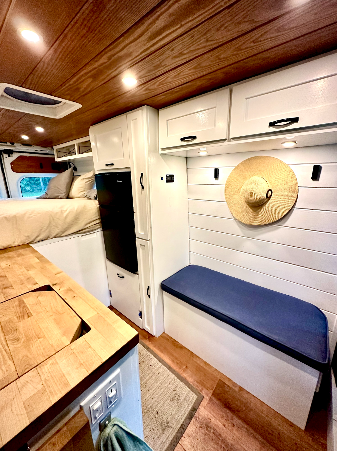 Modern camper van interior: bed, butcher block kitchen, fridge, white cabinets, blue bench. Perfect for an RV rental adventure.